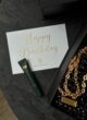 CRYSTAL GLAM: листівка Happy Birthday From the Box, зелений тюбик і ланцюжок сумочки на столі