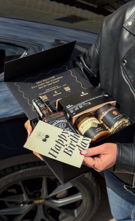 Бокс BOSS: жінка в шкіряній куртці біля авто тримає чорну коробку з Jack Daniel's, кавою та листівкою Happy Birthday