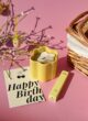 CALM & CARE: жовта свічка-квітка, блиск Lipss і листівка Happy Birthday на рожевому тлі