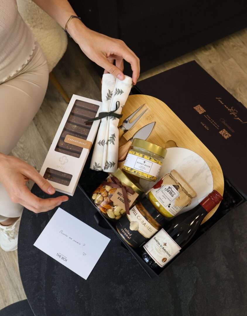 Бокс CHEESE LOVER: жінка розпаковує чорну коробку з бамбуковою дошкою, ножами для сиру, медом, горіхами та вином